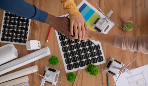 Texte alternatif : Des mains diverses unies sur un bureau avec des panneaux solaires, des modèles d'éoliennes et des matériaux écologiques, symbolisant le travail d'équipe dans des solutions énergétiques durables.