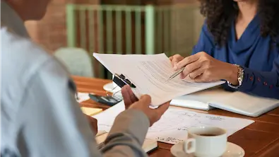 Two professionals discuss a document at a wooden table, surrounded by papers and coffee. One gestures with a pen, creating a collaborative atmosphere.