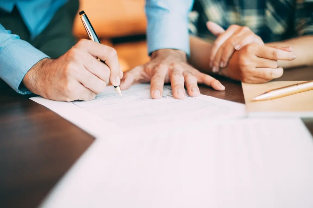Gros plan sur deux personnes examinant un document. Une main pointe, l'autre tient un stylo, prête à signer. La scène évoque la collaboration.