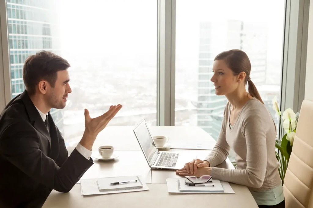 Un homme en costume et une femme en pull discutent amicalement à une table de bureau, avec un ordinateur portable, des tasses à café et des documents. De grandes fenêtres offrent un panorama urbain.
