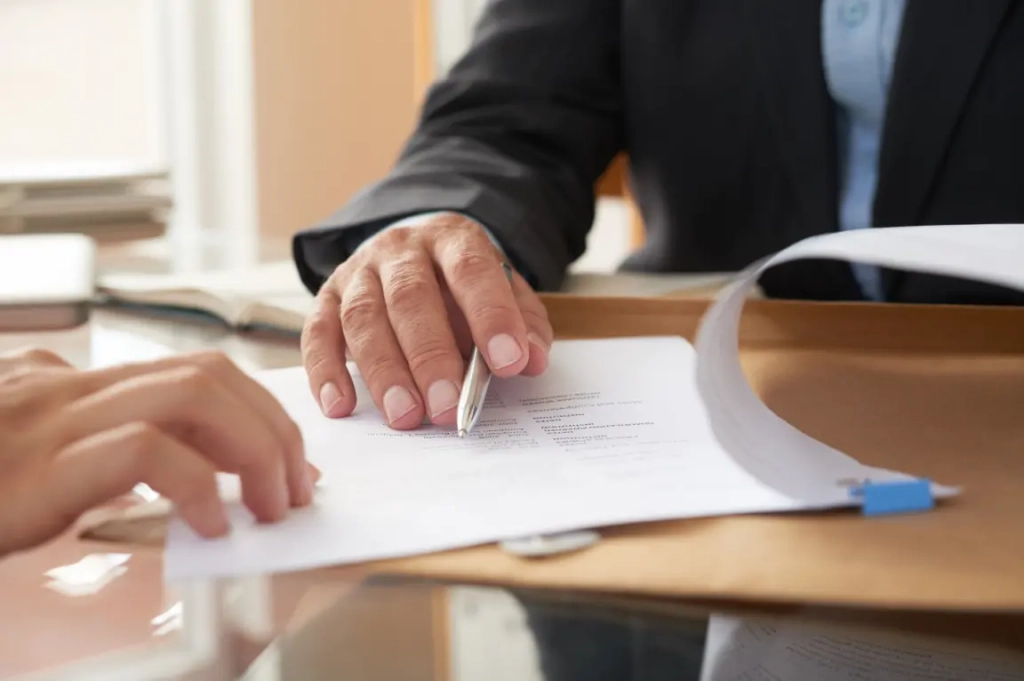 Gros plan d'une table de réunion d'affaires avec une personne en costume pointant un document avec un stylo. Une autre main est visible, suggérant une discussion.