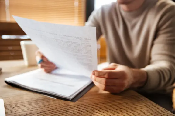 Une personne vêtue d'un pull beige tient et examine des documents imprimés à un bureau en bois, avec une douce lumière naturelle provenant des stores créant une atmosphère calme.