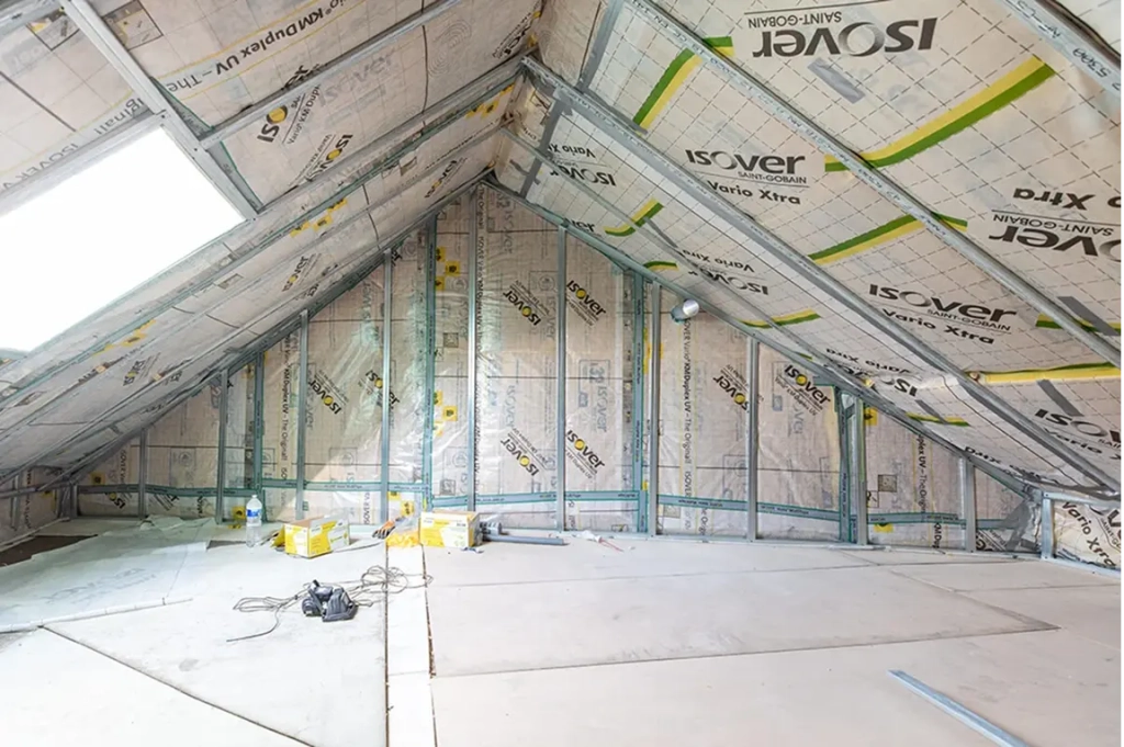 Un grenier en construction, avec isolation et charpente métallique visibles le long du plafond incliné. Des boîtes, des outils et des fils électriques sont éparpillés au sol.