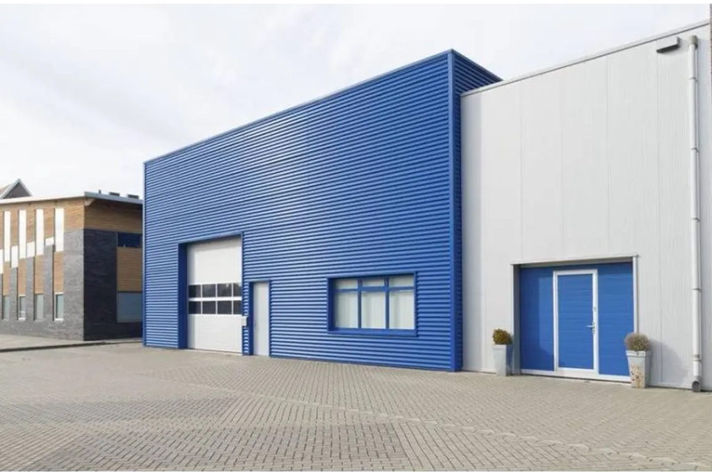 Bâtiment industriel moderne avec façade en tôle ondulée bleue et blanche. Il comprend une grande porte de garage, des fenêtres et des plantes en pot sur une surface pavée.
