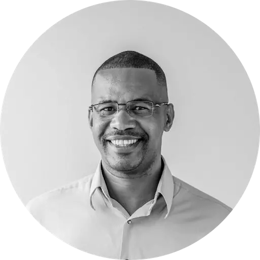 Un homme portant des lunettes sourit dans un portrait en noir et blanc. Il porte une chemise boutonnée de couleur claire sur un fond uni, exprimant une ambiance conviviale.